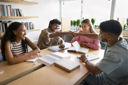 Diverse group of motivated students engaged in productive learning in library seated around desk with open books and notebooks, focus on two guys giving each other fist bump, symbol of teamwork, unity - Powered by Adobe