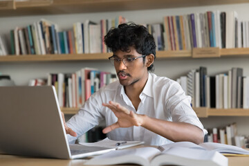 Young Indian male student engaging in videocall, virtual meeting in college library, participate in remote studying, videoconferencing, seminar. Modern distance education, interactive online learning