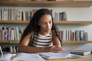 African student girl learning subject, sits at table in public or college library, looks focused,...
