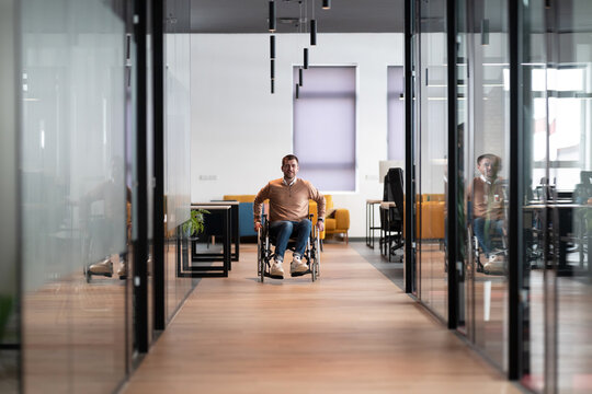 Modern startup coworking office corridor with glass walls, featuring a man in a wheelchair. The space showcases an inclusive, accessible workplace with a sleek, open design and bright lighting.