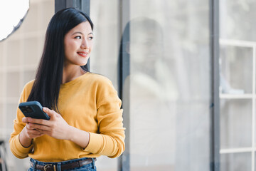 Obraz premium Woman in yellow sweater holding smartphone and smiling while standing near glass window.
