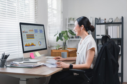 Asian businesswoman working at desk with computer displaying data charts in bright office. - Powered by Adobe