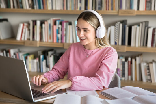 Young focused student woman wear wireless headphones studying online using laptop sit at desk in library, gain new knowledge through modern educational platform. Generation Z, learning, development