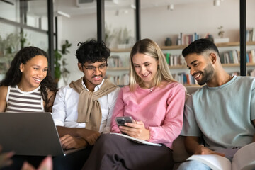 Young Caucasian schoolgirl using her smartphone seated on sofa with diverse schoolmates in high school or university library during break. Social media usage, modern tech overuse, educational content