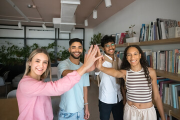 Four young 18s enthusiastic students smile, look at camera stacking their palms together as symbol of joint task completion, university admission celebration, showing team spirit, expressing support