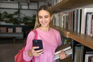 Woman standing in library, holding books and smartphone with contented smile, chatting, or check messages, read text or respond to friend, looks up academic resources, articles, references for studies
