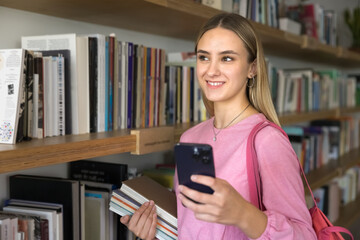 Pensive smiling student girl using smartphone while stand in library, holding workbooks, share messages, check list of literature to choose or school classes using modern mobile application schedule