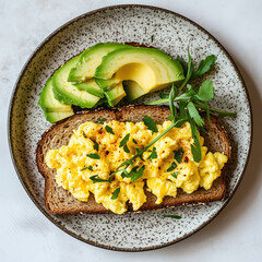 The image shows a plate with a slice of whole grain toast topped with scrambled eggs, garnished with chopped herbs and a sprinkle of pepper. Next to the toast, there are slices of avocado and a sprig 