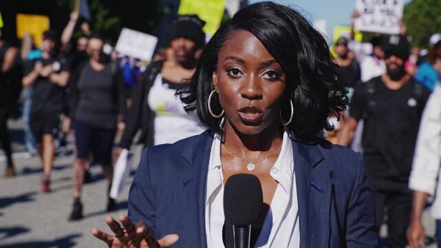 A video frame shows a reporter in a medium close-up angle, speaking into a microphone with a protest crowd blurred in the background.