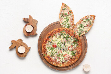 Board with tasty pizza with Easter bunny ears, candles and wooden bunnies on white background