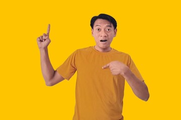 Fototapeta premium A young Asian man in a yellow shirt looks surprised, pointing enthusiastically at something off-frame against a yellow background. The image captures an expressive moment of amazement