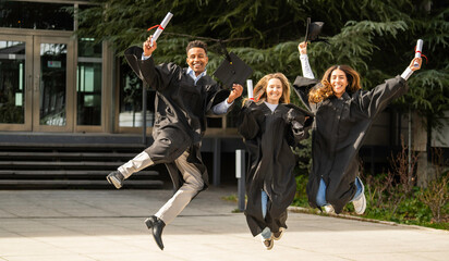 Group of cheerful multiethnic graduates jumping for joy in front of university building,...