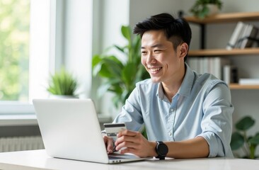 An Asian man is sitting at a table in front of a computer with a bank card in his hands, paying for an online purchase. A bargain, a sale
