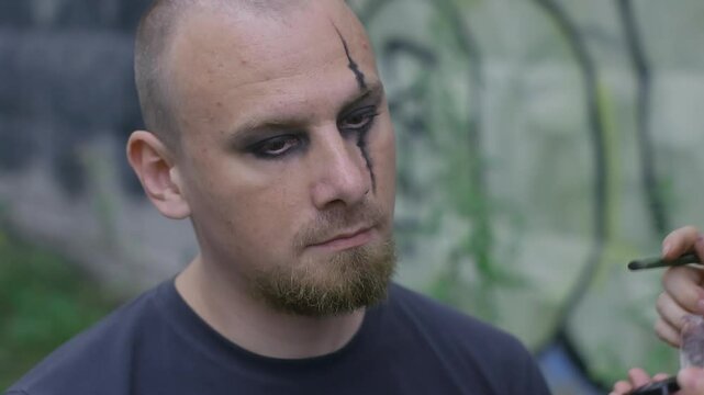 Woman expertly paints elaborate patterns on a bald gothic man's face. He stays motionless as she perfects the look with a brush for Halloween. They get ready for the occasion in an open area