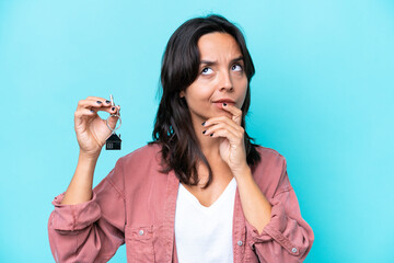 Young hispanic woman holding home keys isolated on blue background having doubts while looking up