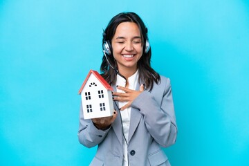 Real estate hispanic agent holding a toy house isolated on blue background smiling a lot