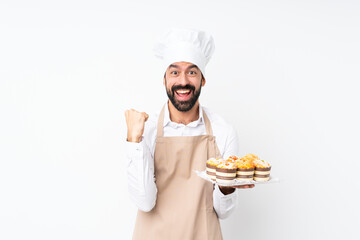 Young man holding muffin cake over isolated white background celebrating a victory in winner position