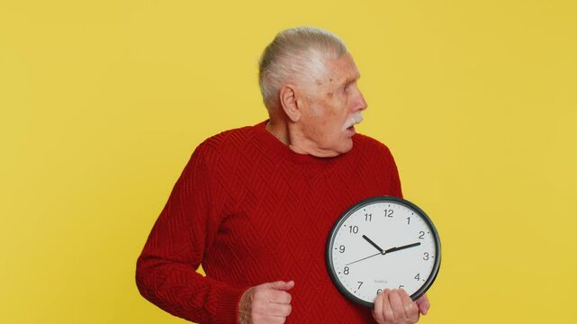 Senior old man with anxiety checking time on clock, running late to work, being in delay, deadline. Elderly grandfather looking at hour, minutes, worrying to be punctual isolated on yellow background