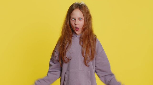 Cheerful funny bully young cute girl showing tongue making faces at camera, fooling around, joking, aping with silly face, teasing. Preteen redhead child kid on yellow background. Little children