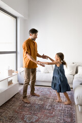 Fototapeta premium Loving mature dad and little, adorable daughter dancing together at home, listening music, holding hands, enjoy active weekend leisure in modern living room, full-length vertical shot. Family pastime