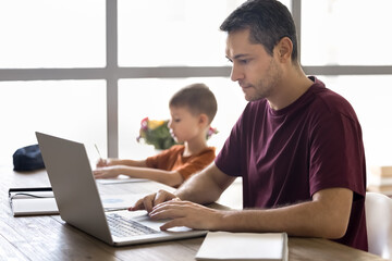 Young 35s focused man working on modern laptop while his little 7s school-age son drawing pictures, study or makes exercise seated together at table at home. Remote work, education, family lifestyle