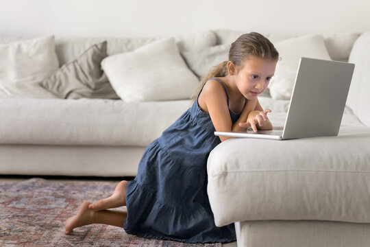 Alpha gen kid girl leaning towards laptop placed on sofa, spend pastime alone on internet, looks focused while websurfing information online, watching educational videos or playing games. Tech overuse
