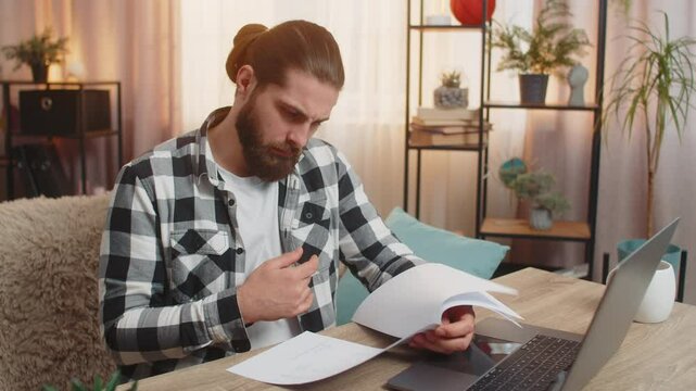 Young serious man checks papers at home table reading fine print new agreement with deep concern studying analyzes contract with a bank, clutches head at complex conditions clauses feeling pressured.