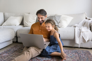 Dad and little daughter enjoy laptop usage seated on floor in cozy living room on weekend at home. Family watching educational, developmental content, make purchase, exploring new creative AI program