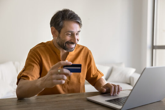 Smiling Portuguese 40s man doing online shopping at home, sit at table with laptop, hold debit or credit card, buying gifts, spend money, making quick transactions. Cashless payments via marketplace