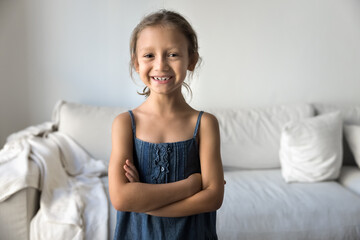 Cute little girl dressed in denim summer dress standing alone in living room with her arms-crossed on chest, looks confident and self-assured, exuding positivity and happiness. Alpha gen kid portrait