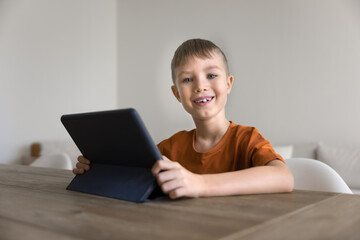 Portrait of cute little boy sitting at table with tablet, look at camera with contented smile, enjoy internet usage for fun or study on leisure at home. Younger gen addicted with gadgets, apps usage
