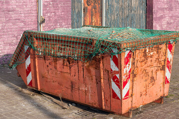 red weathered steel industrial waste container covered with a green net stands outside in front of...