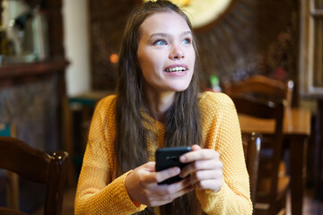 A Cheerful Young Woman is Engaged with Her Smartphone While Relaxing in a Cozy Cafe Setting