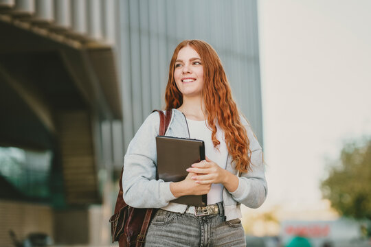 A young woman with a joyful smile is outdoors, holding a tablet and wearing a stylish backpack