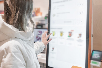 A woman in a white jacket chooses drinks and orders food for dessert in a restaurant or pizzeria coffee shop through a touchscreen screen on her own. The restaurant menu is displayed on the screen in