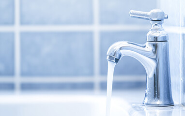 A close-up of a shiny faucet with water flowing, set against a soft blue tiled background, highlighting cleanliness and modern design.