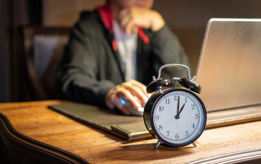 Alarm clock sits on a wooden desk, showing the time, while a businessman in a dark hoodie works on...