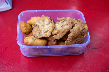 Bakwan (Indonesian vegetable fritters) in a clear plastic container. Overhead shot. Casual street food snack on a red table