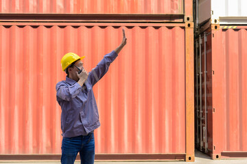 Male dock worker in yellow hard hat and uniform directing operations in front of orange shipping containers by using walkie talkie while raising arm to signal for container loading in shipping cargo y