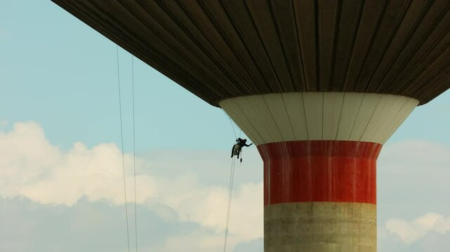 Acrobatic building worker performing restoration on an old brutalist aqueduct suspended by ropes with safety harness featuring industrial architecture construction techniques structural maintenance