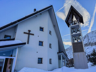 A catholic church with a bell tower in snowy Passo Del Tonale Italy. Clear sunny blue sky with clouds in interesting shapes in Passo Tonale near Italian alps ski resorts. Winter ski travel destination