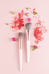 Two chic makeup brushes on a pink delicate background with scattered blush and small flowers. Makeup and beauty concept. The top vertical view.
