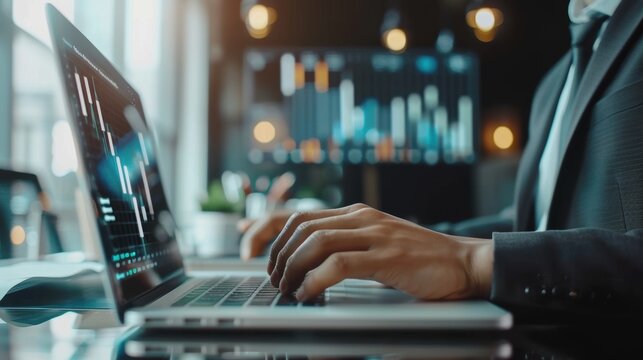 Man is typing on a laptop in front of a monitor with graphs on it