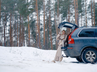 Happy young woman sitting in opened car trunk and drinking hot tea or coffee in snowy forest.