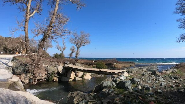  A scenic rustic landscape on Samothrace Island, Greece, featuring a small wooden bridge over a stream leading to the sea. The area is surrounded by rocks, dry trees, and a beautiful coastal view. Goa