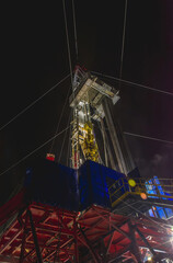 Illuminated industrial drilling rig towers over snowy landscape at night, with operational equipment and pipes creating dramatic silhouette against dark sky