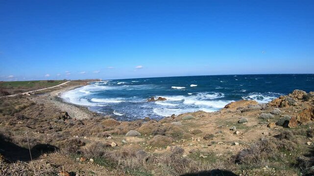 waves crash against a rugged, rocky shoreline under a clear blue sky. The white foam contrasts with the deep blue sea, creating a dynamic coastal scene.