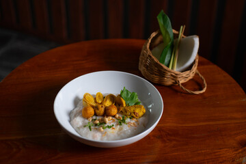 Bubur ayam, a traditional Indonesian food. Rice porridge with sliced chicken, rolled omelette, peanut, sambal (chili sauce), and kerupuk (crackers), poured with curry sauce. Served on wooden table