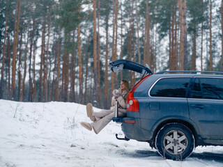 Fototapeta premium Happy young woman sitting in opened car trunk and drinking hot tea or coffee in snowy forest.