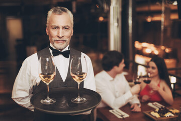 Sommelier is holding tray with glasses of white wine, standing near table of couple who are on date.